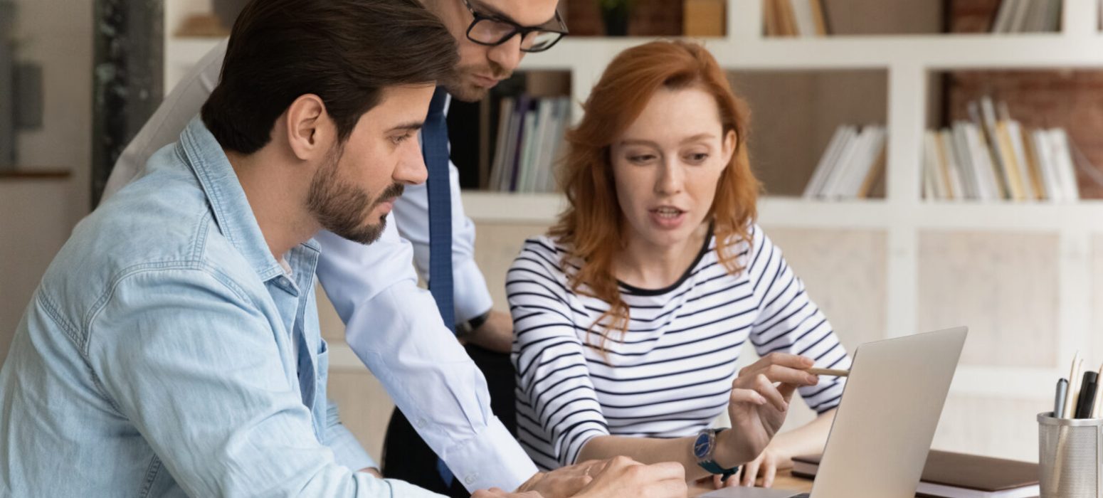 Smart young male leader supervisor reviewing online sales report on computer, working with motivated colleagues. Concentrated three millennial employees preparing presentation in modern office.